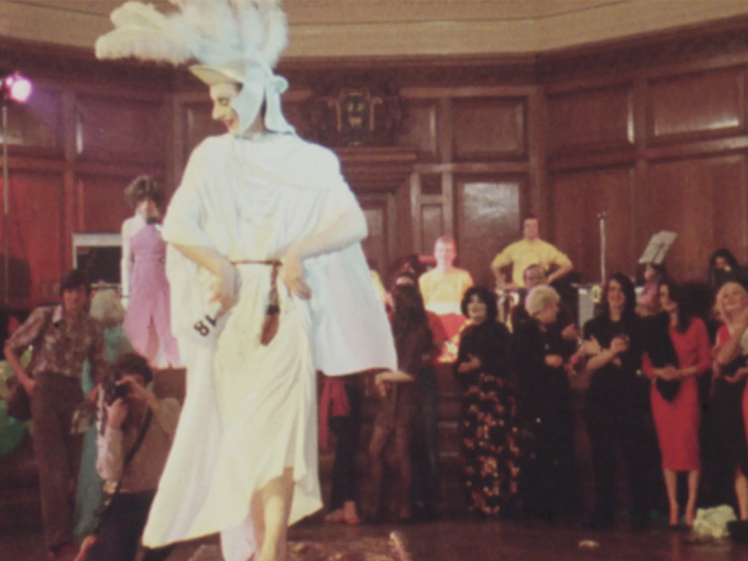 A performer in a white gown and feathered headdress walks before an audience, from Drag Ball.