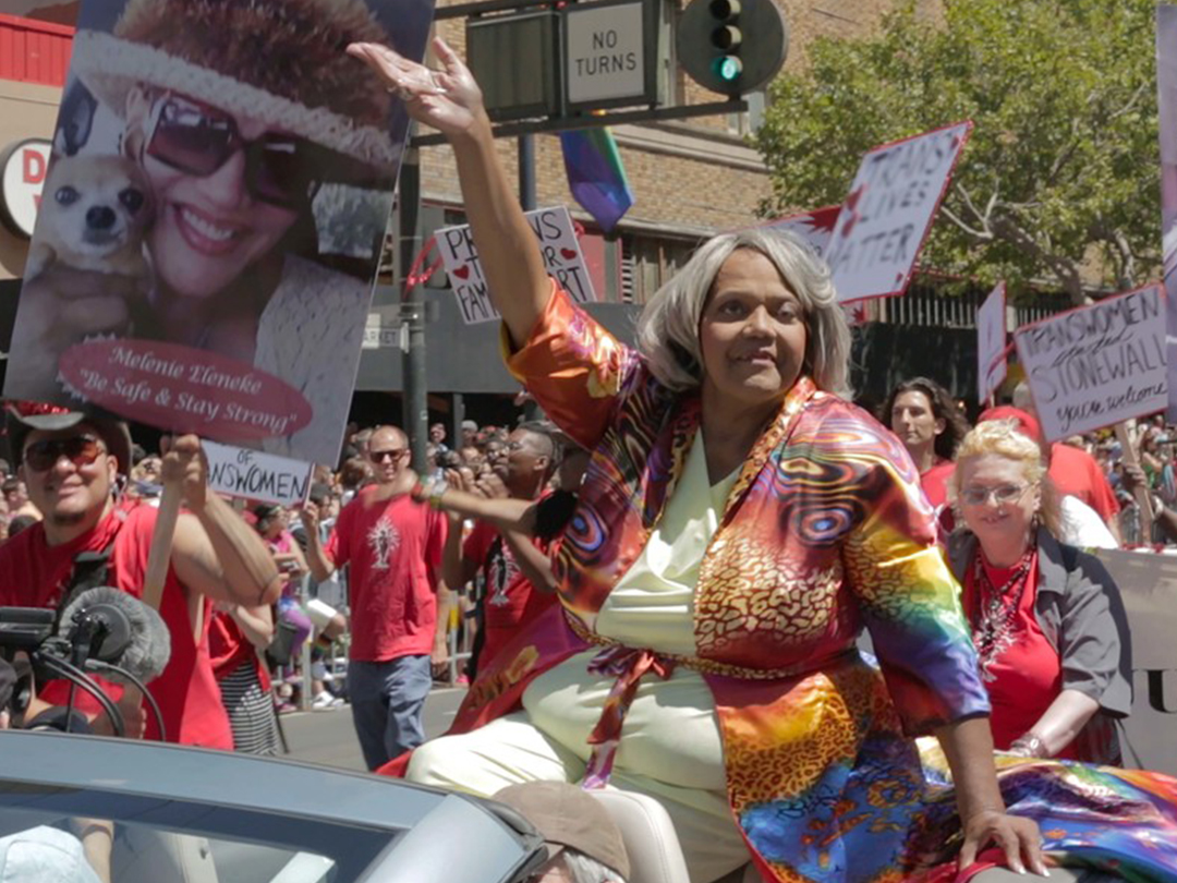 A parade participant waves from an open-top car amid a crowd and placards, from MAJOR!.